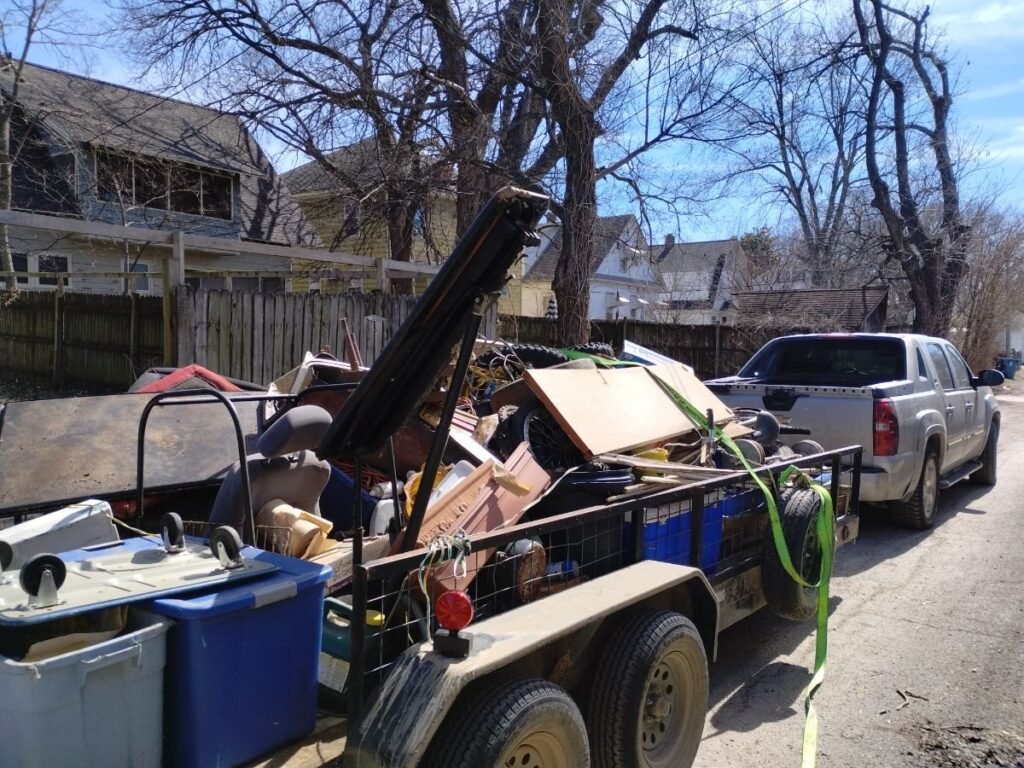 A utility trailer overloaded with miscellaneous junk and debris, including wood, furniture pieces, bins, and metal objects